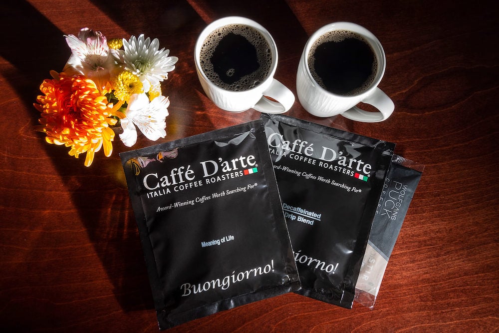 Flatlay on dark, wood table with two mugs filled with coffee, individually packaged coffee sachets and flowers